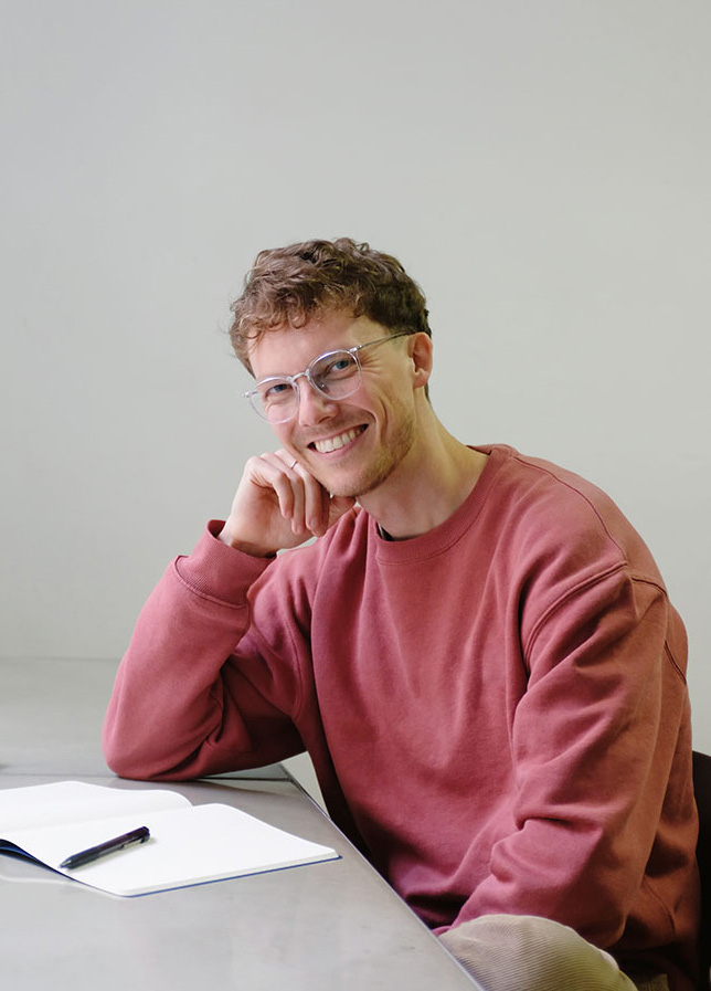 Dr. med. Lukas Möschter im roten Pullover, sitzt am Schreibtisch und schaut freundlich in die Kamera